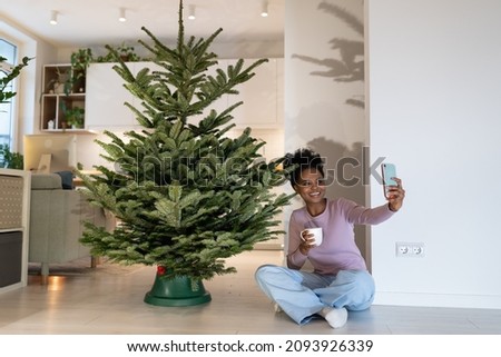 Young woman taking selfie photo sitting at undecorated natural christmas tree enjoy natural beauty and freshness of pine in new apartment. Happy african american female preparing for new year and xmas