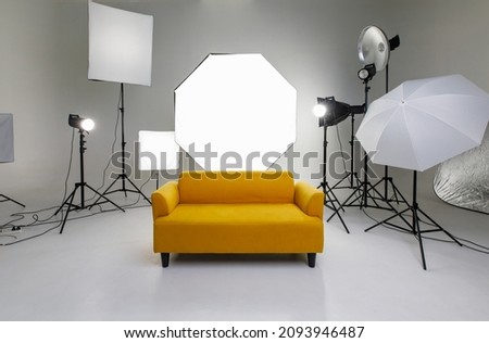 Studio shot fashion backstage photographing shooting set with yellow cozy sofa couch and photography equipment softbox flash strobe spotlight tripods reflector umbrella on white backdrop background. Royalty-Free Stock Photo #2093946487