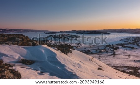Early winter morning in Siberia. The sky is highlighted in orange. Sun glare on a frozen lake. In the foreground is a rocky hill covered with sparkling snow. Baikal. Golden Hour Royalty-Free Stock Photo #2093949166