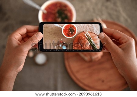 Overhead view of hands holding smartphone and photographing a bowl with freshly cooked Ukrainian traditional soup- Borscht with fresh green raw onion . Mobile hone in live view mode