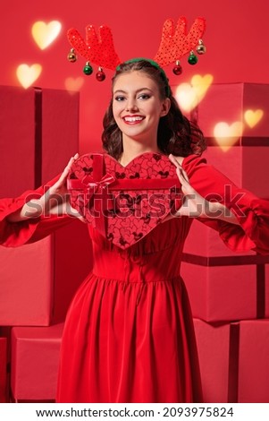 Lovely girl in a bright red dress and deer horns on her head cheerfully holds a heart-shaped gift box. Portrait on a red background with big gift boxes. Christmas and New Year gifts. 