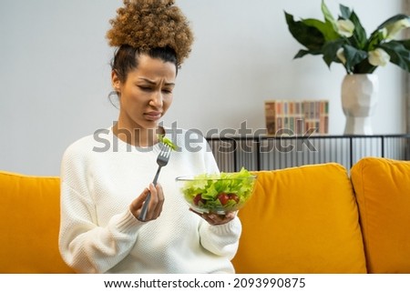 Black woman does not like to eat vegetable salad, healthy food, sitting on the couch Royalty-Free Stock Photo #2093990875