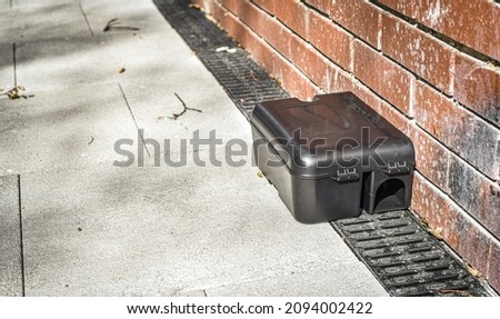 Black rodent bait and trap station being used outside a building to control rats.  Royalty-Free Stock Photo #2094002422
