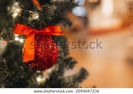 Red Christmas toy with silky bow hanging on the Christmas tree. Magic details with bokeh lights on background. Winter holidays cozy magic details