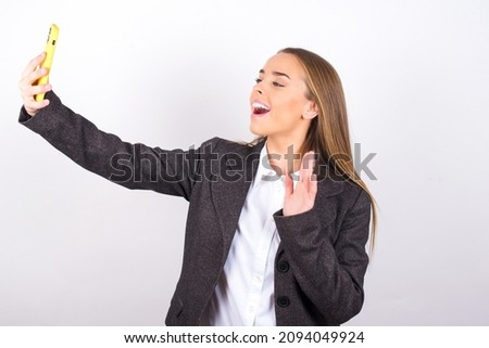 Young business woman wearing jacket over white background holds modern mobile phone and makes video call waves palm in hello gesture. People modern technology concept