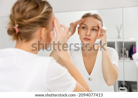 A young beautiful woman looks in the mirror and pulls the skin on her forehead. The view from behind the shoulder, at the reflection in the mirror. The concept of rejuvenation and facial wrinkles. Royalty-Free Stock Photo #2094077413