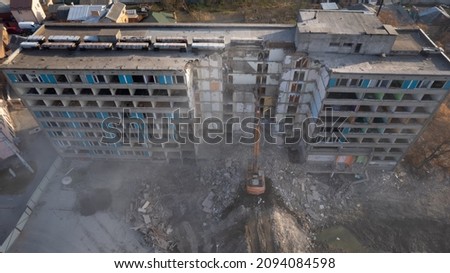 Excavator for dismantling buildings, using a special claw, dismantles a multi-storey building. The sun's rays break through a thick cloud of dust in front of the ruined building Royalty-Free Stock Photo #2094084598