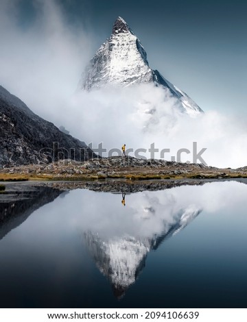 Hiker man walks alone through the incredible Swiss Alps with the Matterhorn in the background between clouds and mystery while it is reflected in the Riffelsee lake Royalty-Free Stock Photo #2094106639