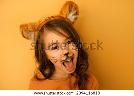 A child dressed up as a tiger with a painted mustache and a hat with ears growls and shows his tongue. Funny cute beautiful girl in the image. lunar cycle of the Chinese animal zodiac. New year 2022 Royalty-Free Stock Photo #2094116818