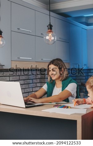 Caucasian young woman working online on laptop with daughter doing her homework in the kitchen. Internet network. Online education.