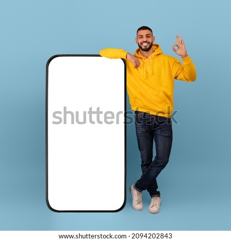 Excited arab man showing okay gesture, standing next to huge smartphone over blue background, empty space for mobile app or website design on screen. Advertisement mockup
