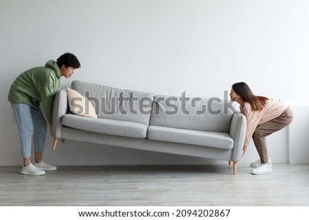 Side view of cheery young Asian couple moving sofa at home, replacing furniture in living room, blank space. Happy millennial spouses making rearrangement in their house together Royalty-Free Stock Photo #2094202867