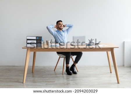 Taking Break. Smiling young Arab man relaxing on chair sitting at table and resting, using pc laptop, happy millennial male leaning back at workplace, enjoying his job, feeling pleased and satisfied Royalty-Free Stock Photo #2094203029