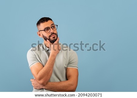 Pensive middle eastern guy thinking and touching chin, looking aside at free space, posing over blue background, studio shot. Let me think concept