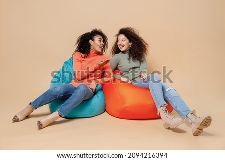 Full size body length two smiling charming young curly black women friends 20s wear casual shirts clothes sit in bag chair speaking talking isolated on plain pastel beige background studio portrait