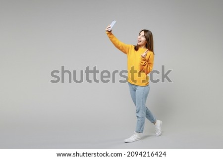 Full length body of young smiling caucasian happy woman 20s in casual knitted yellow sweater doing selfie shot on mobile phone show victory v-sign gesture isolated on grey background studio portrait.