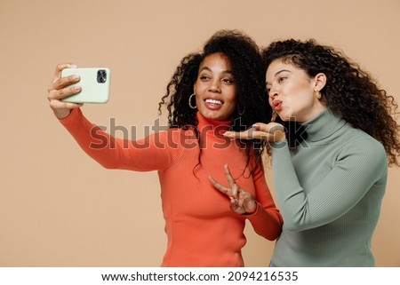Two charming young curly black women friends 20s wear casual shirts clothes doing selfie shot on mobile cell phone blowing sending air kiss isolated on plain pastel beige background studio portrait