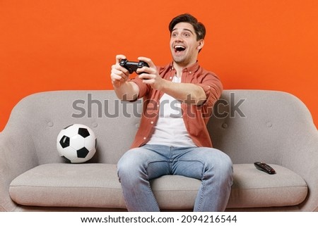 Young happy fun man football fan in shirt support team with soccer ball sit sofa home watch tv live stream play online pc game with joystick console isolated on orange background People sport concept Royalty-Free Stock Photo #2094216544