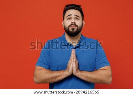 Young pleaded sad caucasian man 20s wear basic blue t-shirt hold hands folded in prayer gesture, begging about something isolated on plain orange background studio portrait. People lifestyle concept