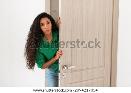 Portrait of confused woman standing in doorway of her apartment, millennial female holding ajar door looking peeking out with frightened facial expression. Unwanted visitor coming without invitation Royalty-Free Stock Photo #2094217054