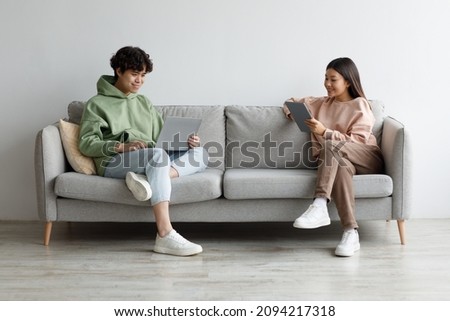 Handsome young Asian guy and his girlfriend using modern gadgets while sitting on couch at home, copy space. Full length of millennial couple communicating online via laptop and tablet pc Royalty-Free Stock Photo #2094217318
