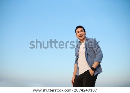 Portrait photo of a happy smiling Asian man wearing a blue t'shirt against a blue sky backround.