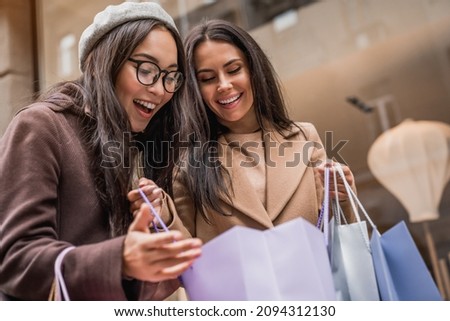 Happy young women looking into shopping bags of her friend near shop showcase outdoor. Buying clothes presents for holidays on sale discount. Black friday Royalty-Free Stock Photo #2094312130