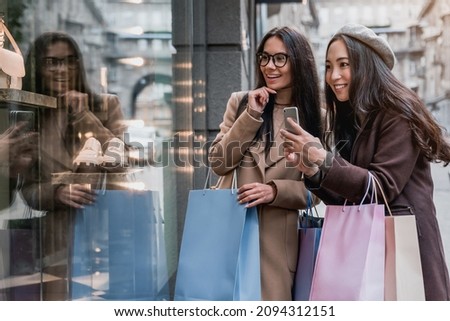 Two happy girlfriends women looking on the shopwindow while photographing on smartphone with shopping bags near the mall. Black friday concept. Discounts and sales