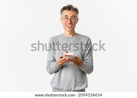 Image of handsome middle-aged man looking surprised and happy, being congratulated with birthday, holding b-day cake and smiling amazed, celebrating over white background