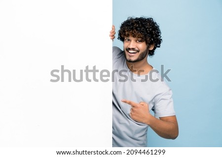 Positive curly-haired Indian or Arabian guy, in casual t-shirt, peeking out from behind advertisement white board, demonstrating blank copy space for your text or design, on isolated blue background Royalty-Free Stock Photo #2094461299