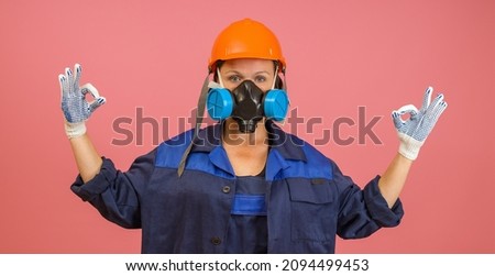 woman in blue uniform, orange helmet, respirator shows ok sign with two hands on pink background