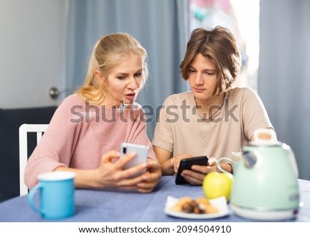 Mother and son sitting at kitchen with smartphones during breakfast and talking