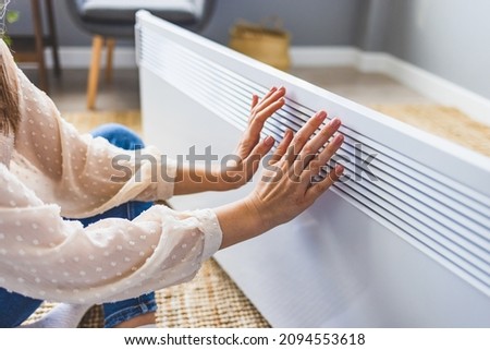 Woman warming hands near electric heater at home. Using heater at home in winter. Woman warming her hands. Heating season. Young woman warming hands near electric heater at home