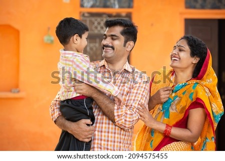 Young happy indian parents holding cute little child having fun and laughing while standing outdoor at village house. Mustache man wearing kurta and woman wear sari. rural india concept. Royalty-Free Stock Photo #2094575455