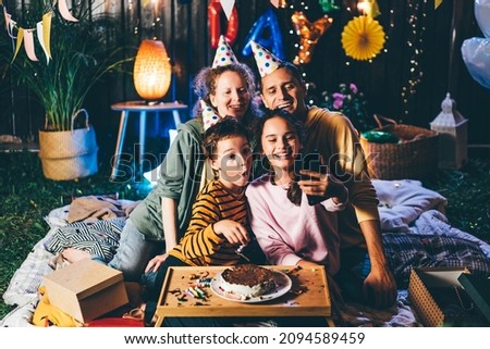 Pretty junior schoolgirl makes selfie with happy family in birthday cones with smartphone at celebration party in decorated cottage yard in evening Royalty-Free Stock Photo #2094589459