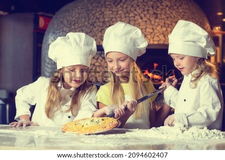 Near the oven, three cheerful little girls, soiled in flour and dressed in chef's uniforms, are making pizza.