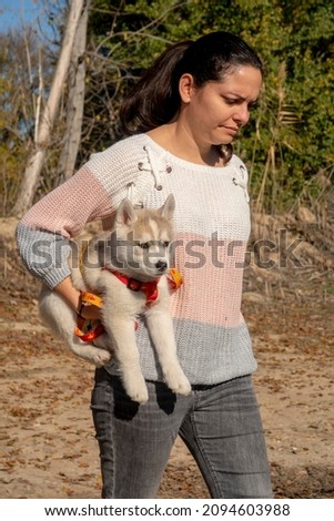 a brunette Latina woman carries her Siberian husky puppy in her arms with the blurred background of several trees in a forest in autumn