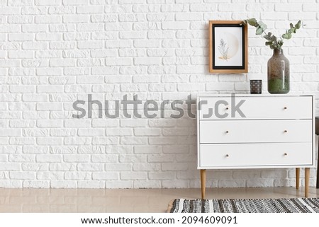 Modern chest of drawers with vase and candle near brick wall
