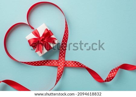 Top view photo of valentine's day decorations white giftbox with red bow in red satin ribbon heart on isolated pastel blue background with empty space