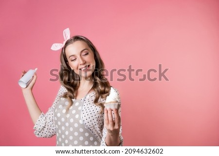 Young charming woman pastry chef and delicious muffin with cream. A cook in a polka-dot apron. pastry chef in an apron on a pink background.