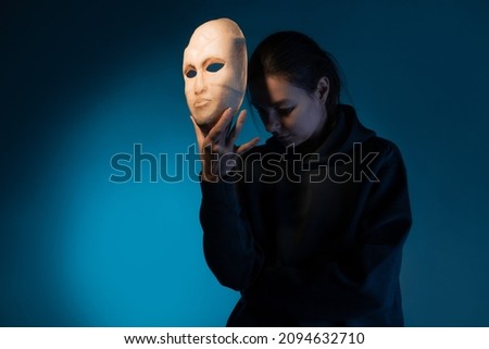 Hiding behind a mask, a young woman in a dark hoodie hides her face with a mask, self-identification problems and impostor syndrome. Portrait in the studio on a dark gray background. Royalty-Free Stock Photo #2094632710