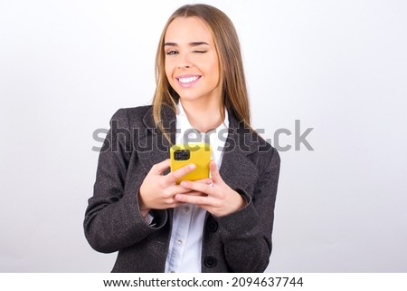 Pleased Young business woman wearing jacket over white background using self phone and looking and winking at the camera. Flirt and coquettish concept.