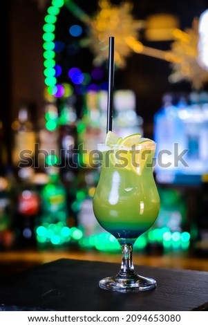 A cocktail in a tall green glass, lemon wedges and a black straw. 
