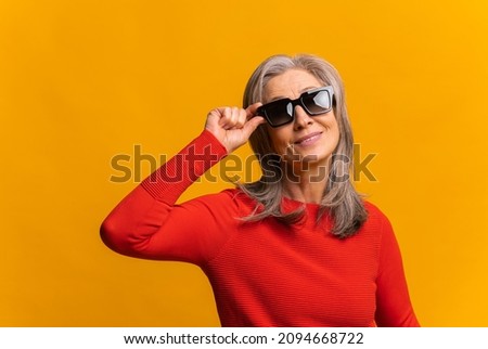 Waist up portrait view of the mature fashionable woman wearing sunglasses posing to the camera over the yellow wall. Stock photo
