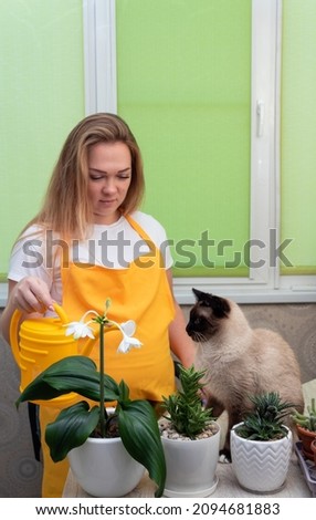 Woman shows watering can to cat sitting next to them, there are potted plants in front of them. Caring for domestic plants and communicating with animals. Selective focus.