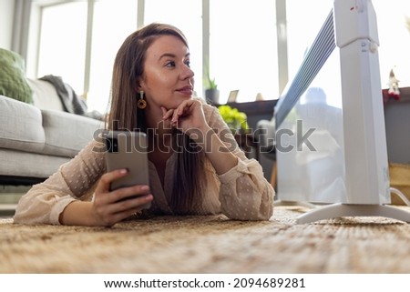 Woman dressed in beige sweater regulating heating temperature with a modern wireless thermostat and smart phone at home. Synchronization of thermostat with mobile devices concept