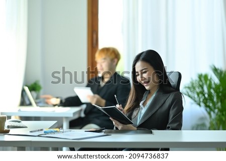Photo of a young businesswoman checking a business information at the working desk over the colleague in the office as a background.