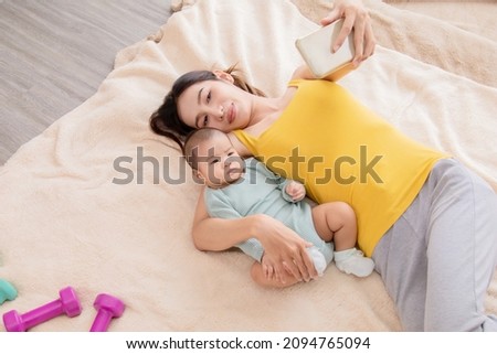 Beautiful Asian mother taking selfie picture by smartphone with daughter in bed, infant lie down on mom arm. Women and baby lie down together and pay attention with camera. 