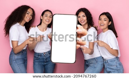 We Recommend. Portrait of smiling multiracial women holding big cell phone white blank device screen in hand, pointing at gadget with free copy space for mock up, banner, pink studio background