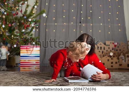 Young happy mom and daughter in red sweaters read books at home. Christmas mood, DIY home decor from cardboard.